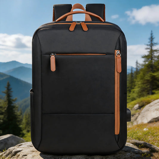 A black backpack with tan leather-like trim and handles, standing on a rock with a mountain and forest landscape in the background.
